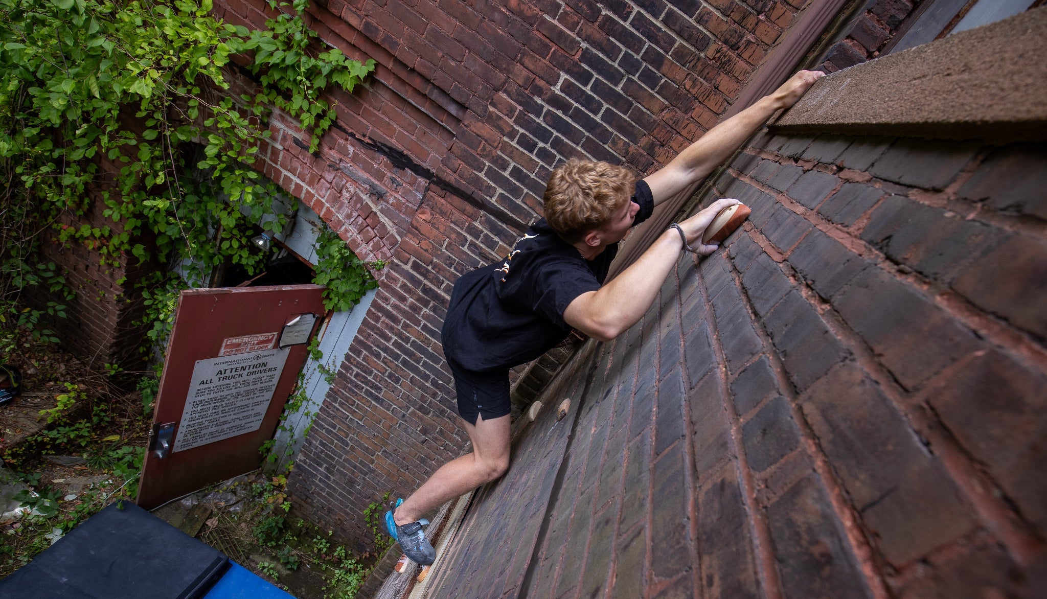 Person climbing a brick wall with a sign and greenery in the background